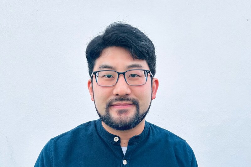 slightly smiling man with thick black hair, glasses, moustache and beard, wearing a blue shirt
